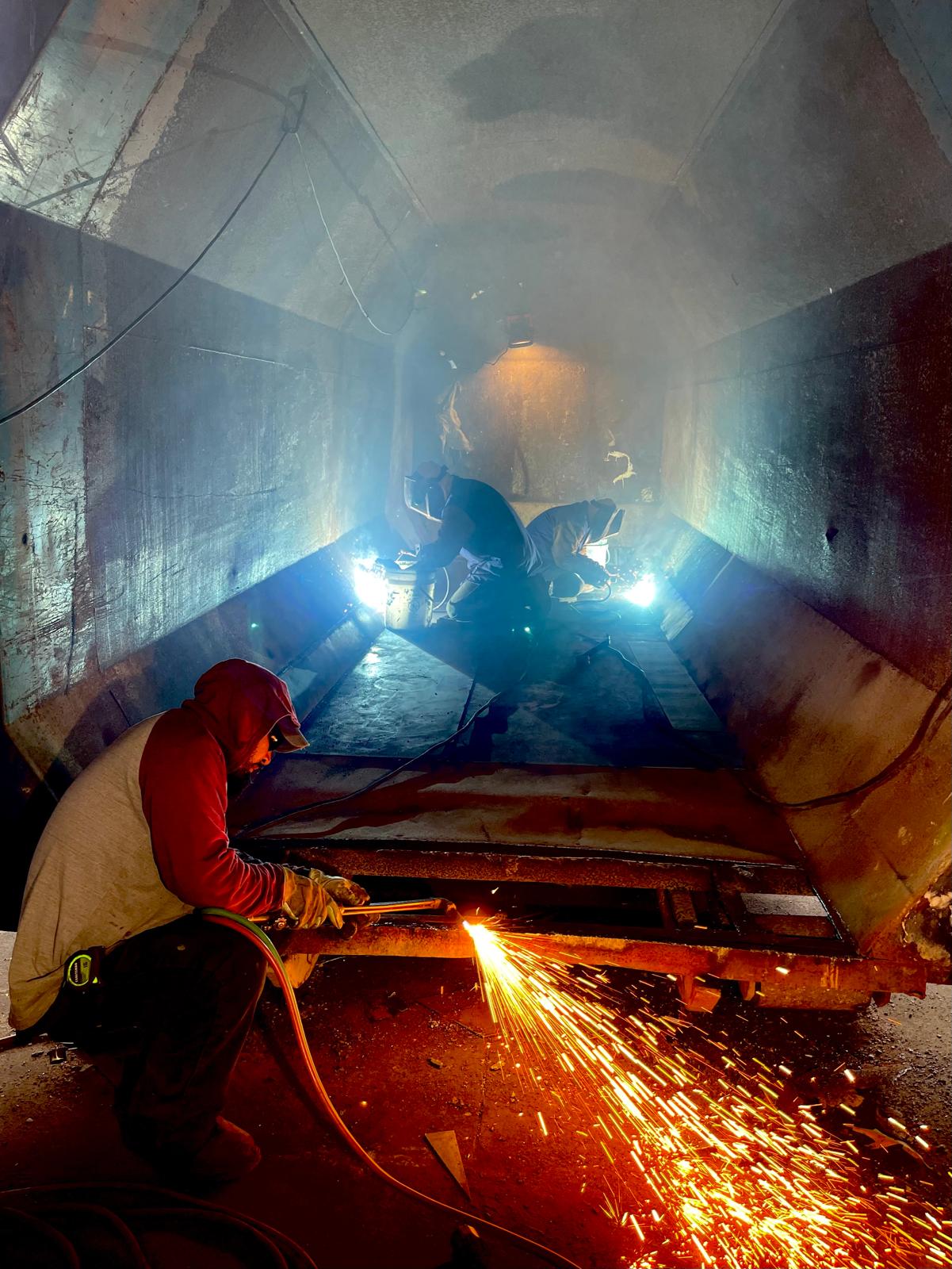 Inside a long steel channel, a worker in a red hooded sweatshirt cuts metal with a torch as orange sparks fall, while two welders in helmets work further back under blue arc light.