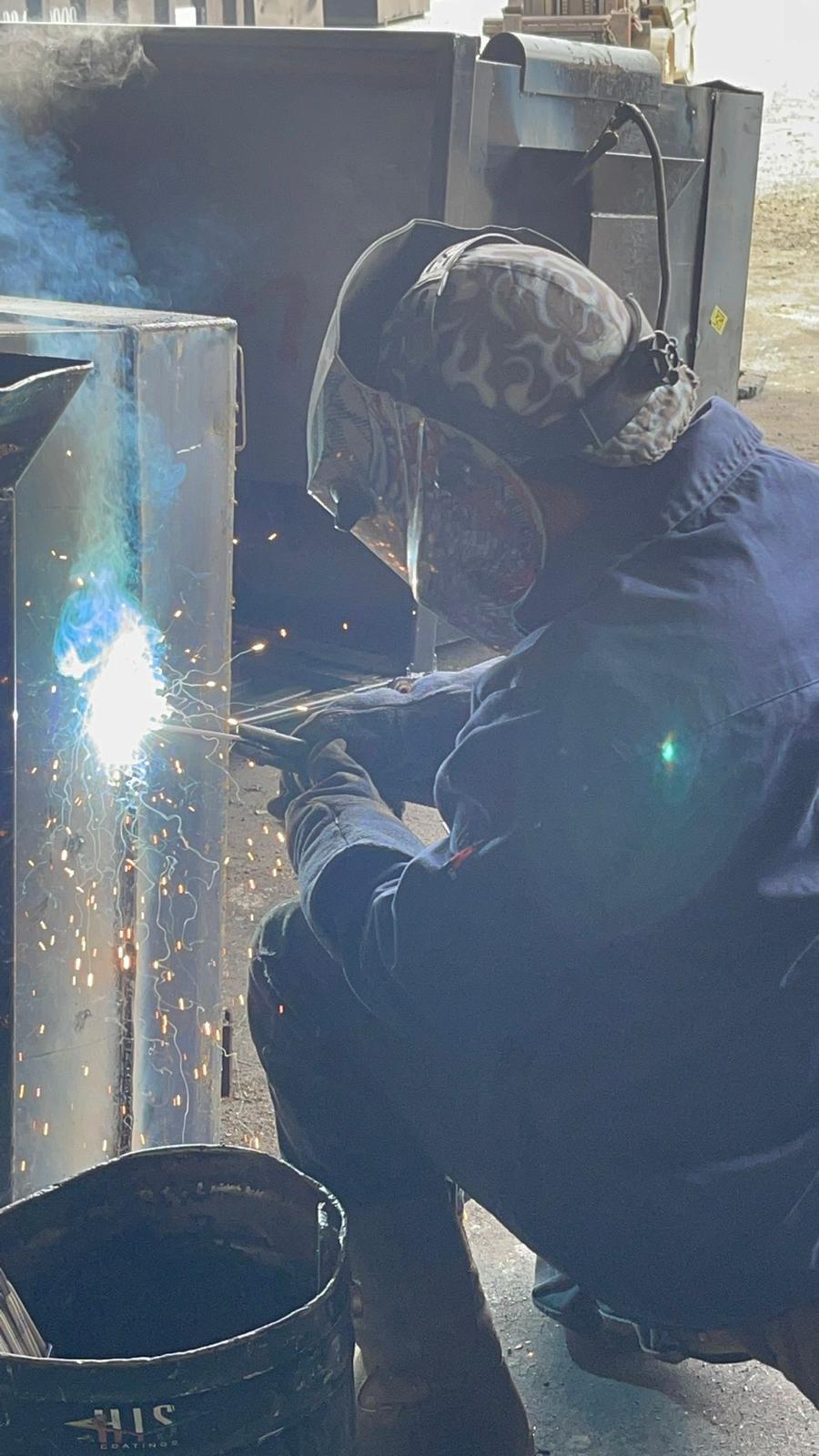 A welder in a patterned cap, welding helmet and navy coveralls crouches to weld a steel cabinet, with sparks and blue light at the seam.