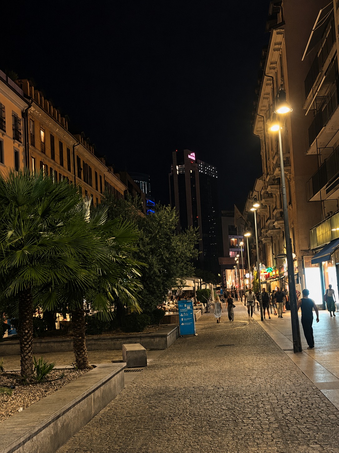 Palm-lined Milan street at night with warm storefront lights and pedestrians.