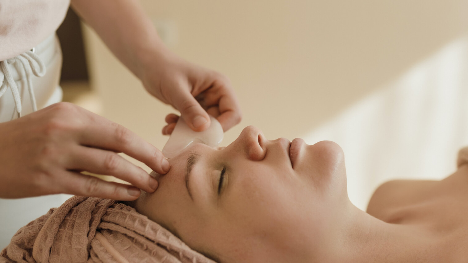 A practitioner drawing a smooth jade gua sha tool along a reclining client's cheek during a facial treatment.
