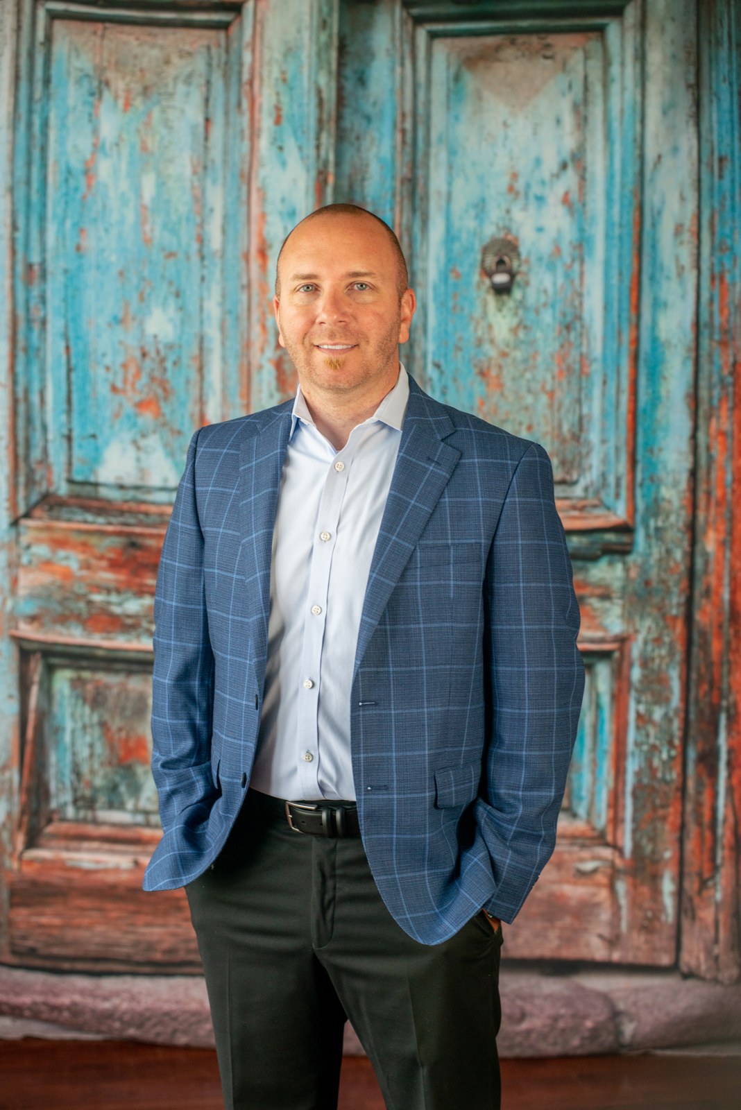 Portrait of Zach Dillard, owner of Edward J. Henry and Sons Auto Body, in a blue windowpane jacket against a weathered turquoise door