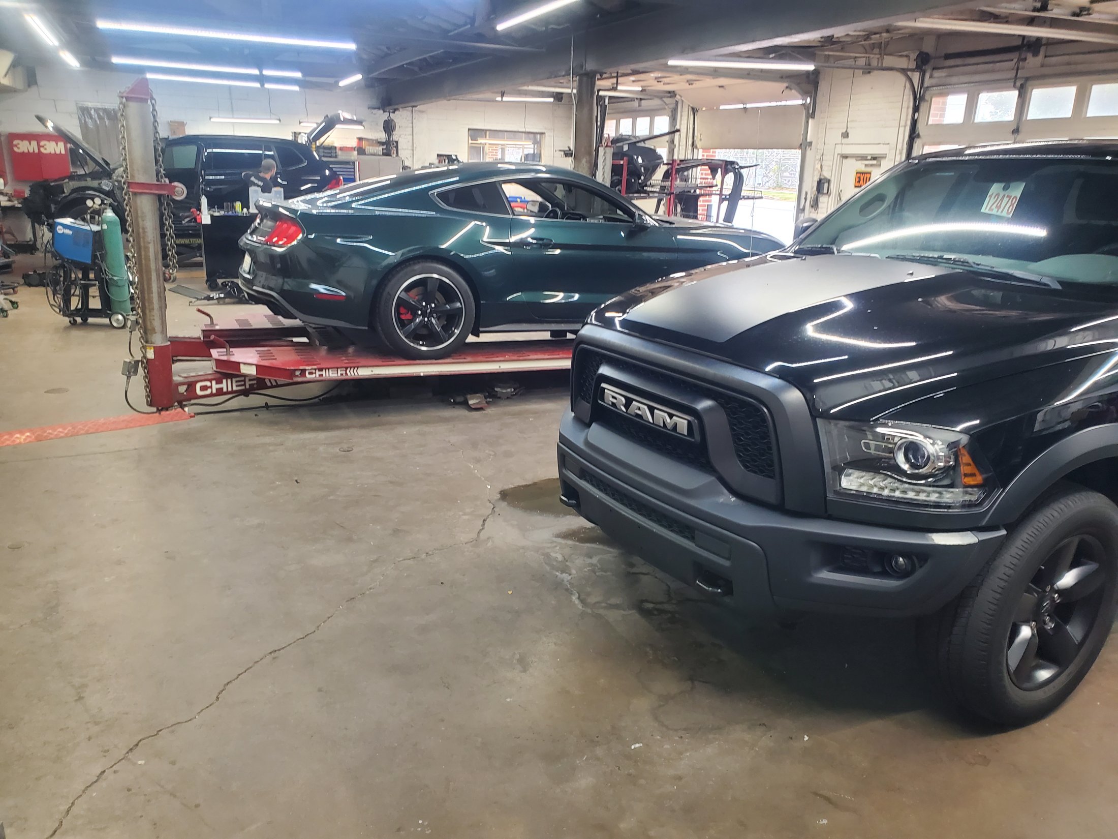 A green Ford Mustang Bullitt on a Chief frame rack in the Henry's shop, a black Ram pickup in the foreground and a technician working in the background