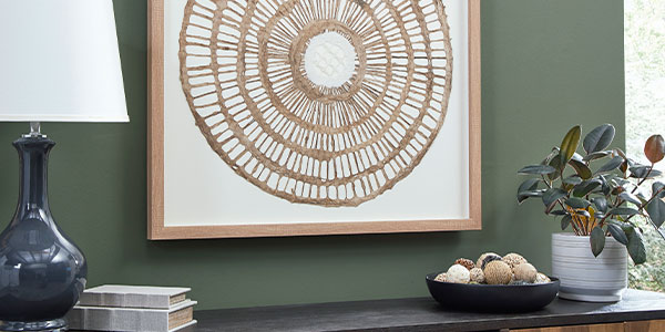 Woven circular wall art, a blue glass lamp and decorative objects against a green wall.