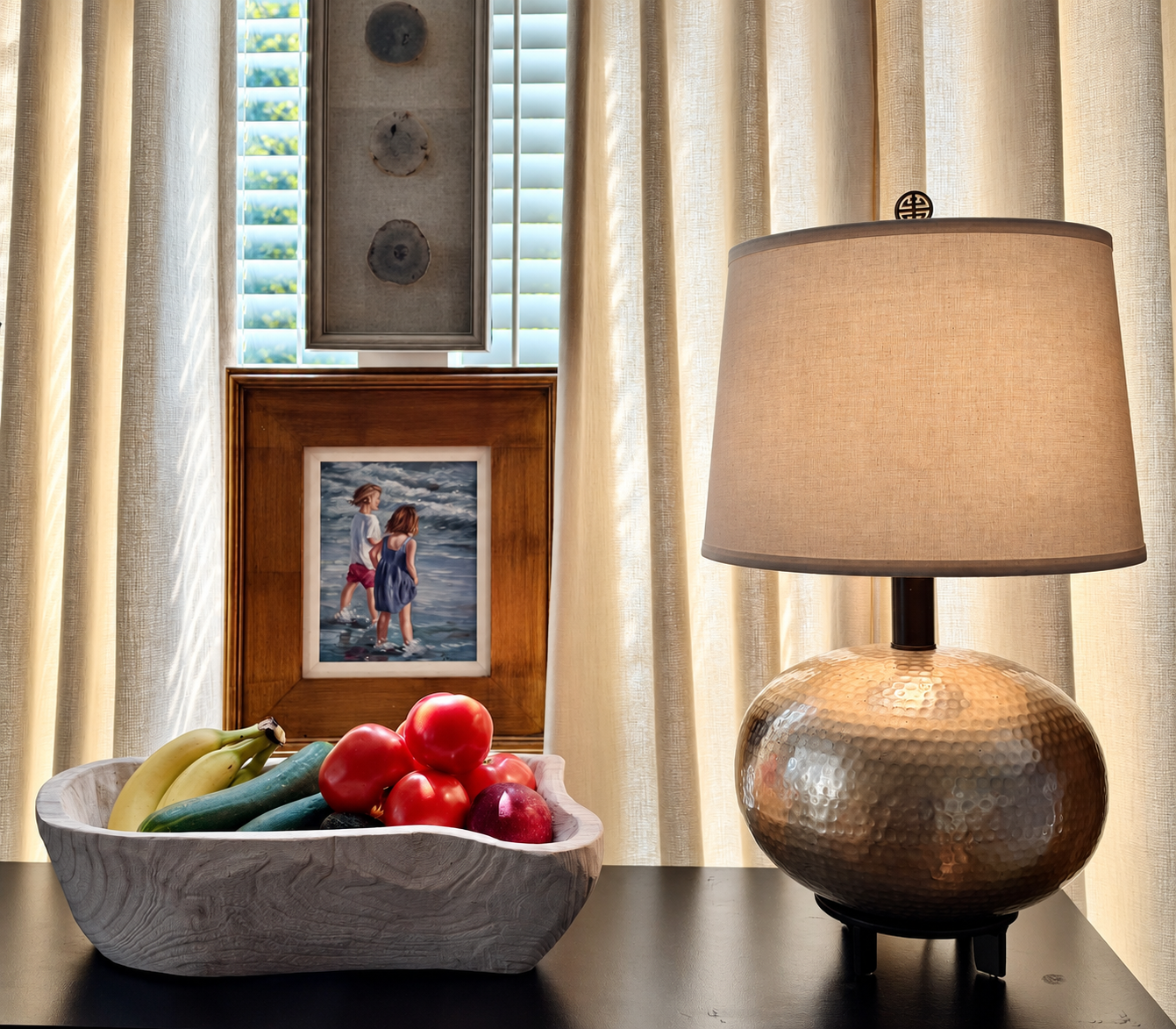 A hammered-metal table lamp with a woven shade beside framed photographs and a carved wood bowl of vegetables.