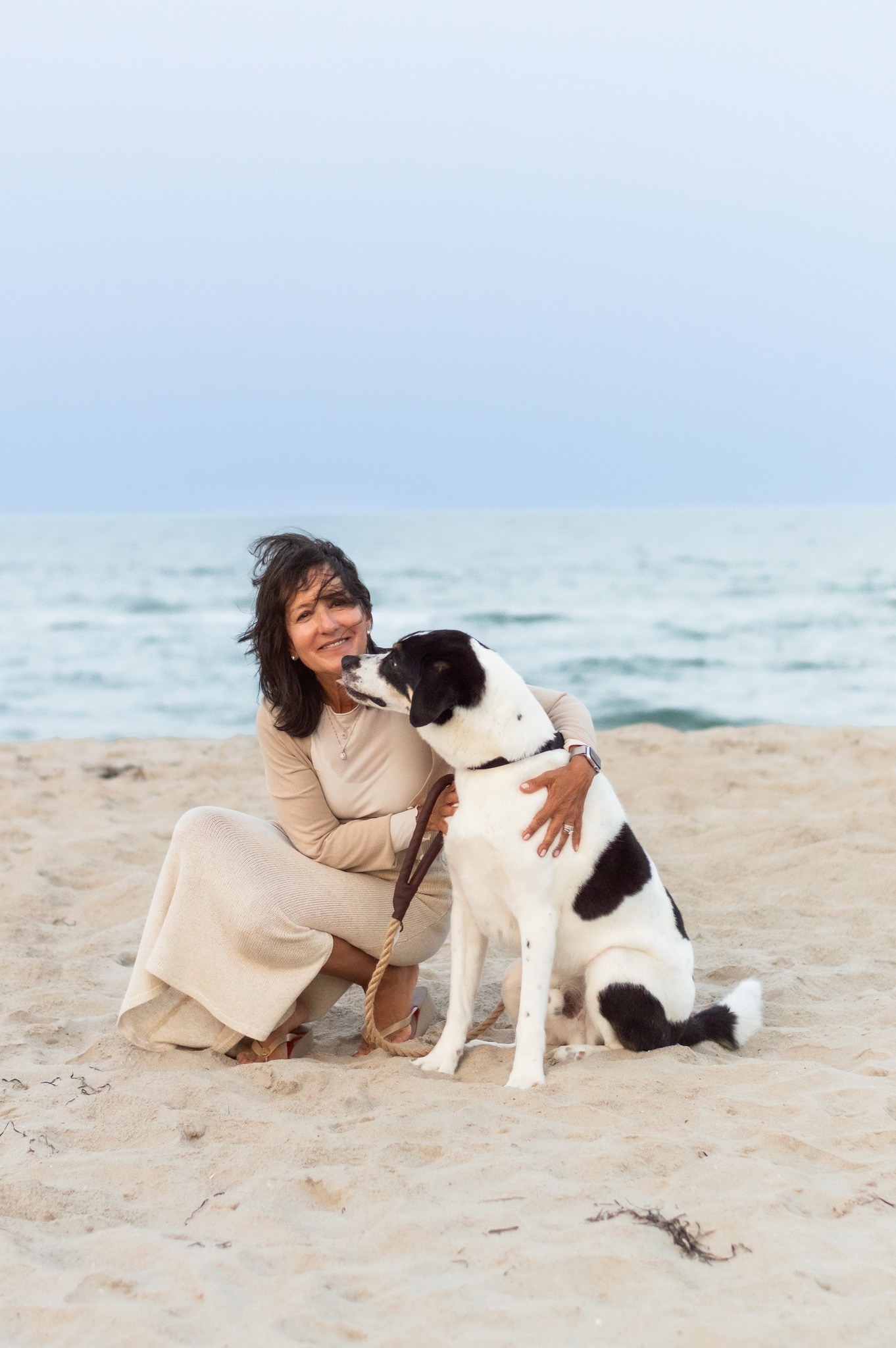 Elaine Orbino seated on a beach beside a black-and-white dog, with the sea behind them.