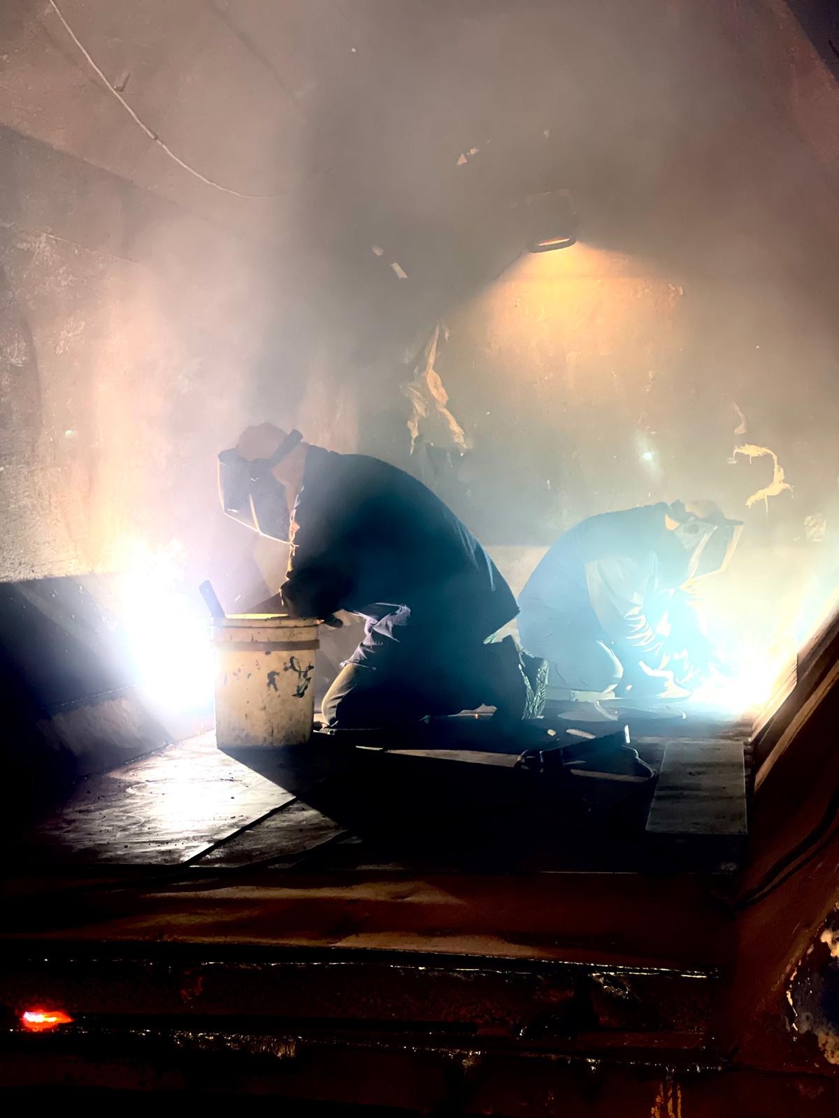 Two welders in hoods and face shields kneel in a haze of smoke, silhouetted by bright blue welding arcs and a single work lamp.