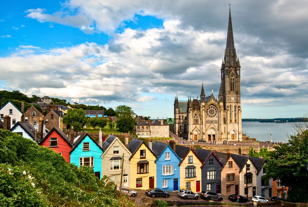 Row of brightly painted houses on a hillside below a tall cathedral beside an Irish harbor.