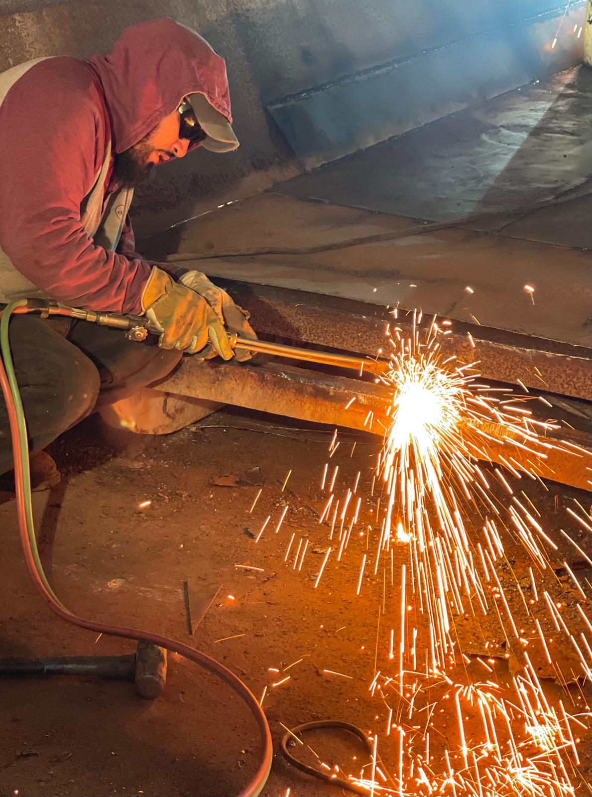 A worker in a red hooded sweatshirt, cap and leather gloves crouches to cut a rusted steel beam with a torch, sending a shower of sparks across the floor.