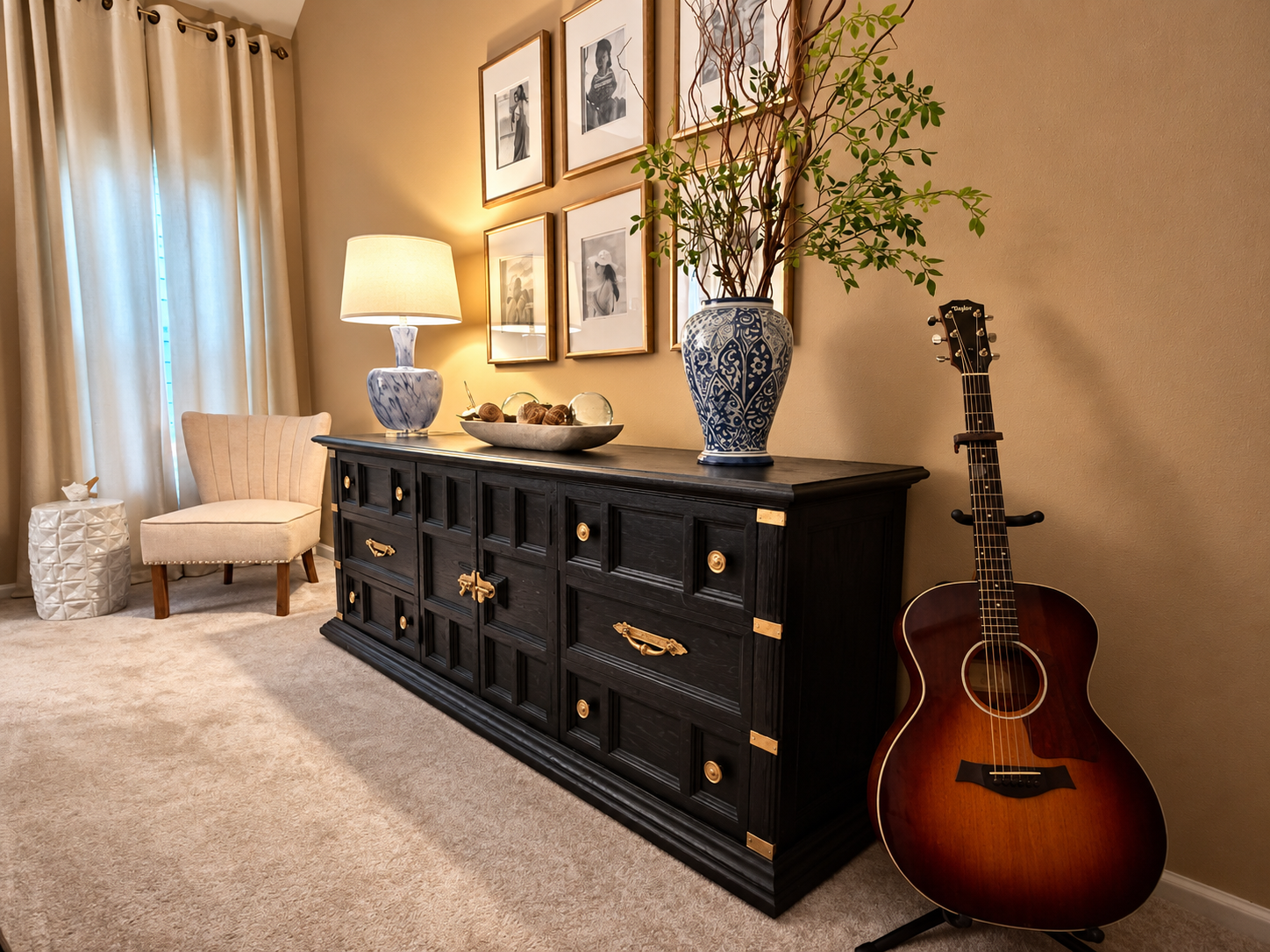 A dark cabinet with brass-tone handles beneath framed photographs, with a vase of branches and a guitar beside it.