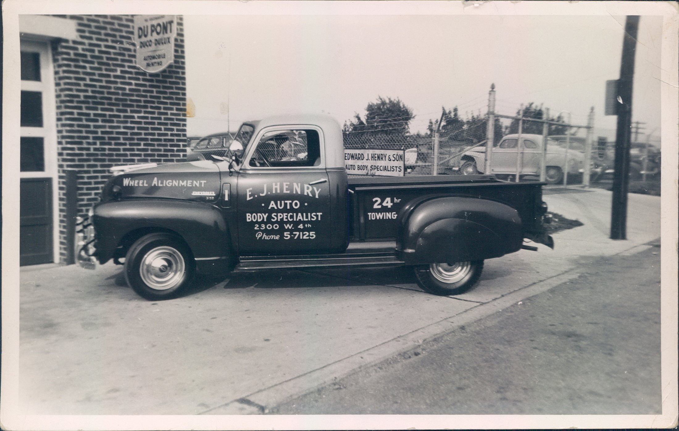 A vintage Chevrolet pickup truck lettered E. J. Henry Auto Body Specialist, 2300 W. 4th, phone 5-7125, 24 hour towing, photographed in the 1950s outside the same building on West Fourth Street