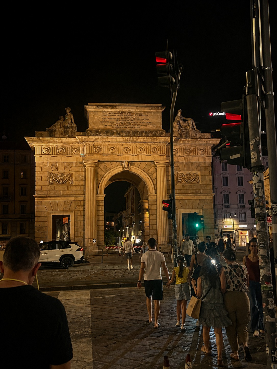 Illuminated historic arch in Milan at night with pedestrians crossing the street.