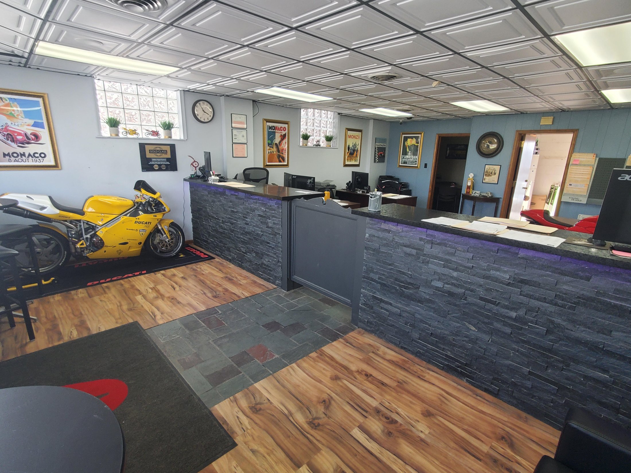 The lobby at Edward J. Henry and Sons, a slate reception counter under Monaco 1937 and Monza racing posters, a yellow Ducati 748 parked on a Ducati rug